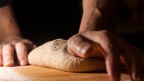Das Bild zeigt zwei Hände, die auf einer bemehlten Fläche einen Brotteig kneten.